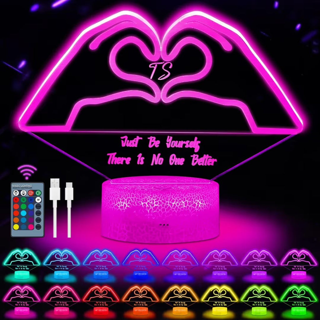 A pink LED heart-shaped night light with a cylindrical base, shown on a black background with a remote control, charging cable, and multiple color options displayed along the bottom. The main product features a heart design with the words 'Just Be Yourself There Is No One Better' and a central 'TS' monogram.