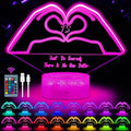 A pink LED heart-shaped night light with a cylindrical base, shown on a black background with a remote control, charging cable, and multiple color options displayed along the bottom. The main product features a heart design with the words 'Just Be Yourself There Is No One Better' and a central 'TS' monogram.