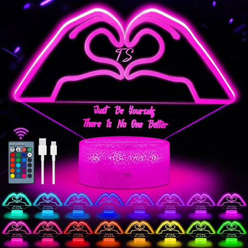 A pink LED heart-shaped night light with a cylindrical base, shown on a black background with a remote control, charging cable, and multiple color options displayed along the bottom. The main product features a heart design with the words 'Just Be Yourself There Is No One Better' and a central 'TS' monogram.