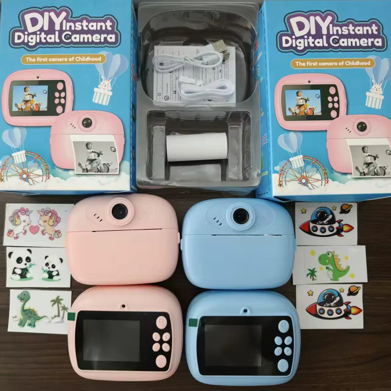 DIY instant digital camera set shown with two pink and two blue cameras, their retail boxes, a plastic roll holder with charging cable, and small printed sticker sheets on a dark wood surface.
