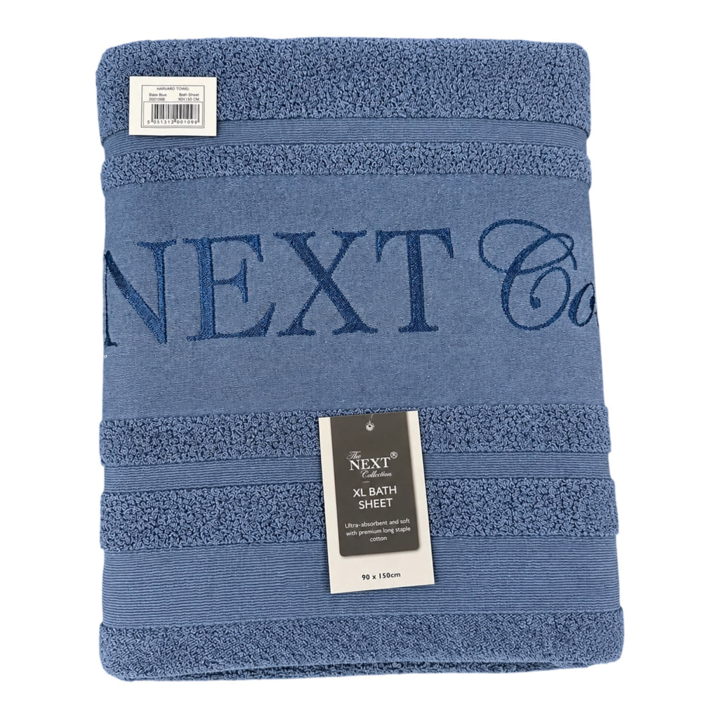A folded blue bath sheet with 'NEXT Co' embroidered across the front, featuring horizontal textured stripes and a hanging product tag.