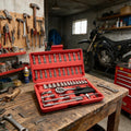A red socket wrench set in an open plastic case sits on a wooden workbench in a garage workshop. Wrenches hang on a pegboard, a motorcycle is parked in the background, and other tools are scattered around the workbench.